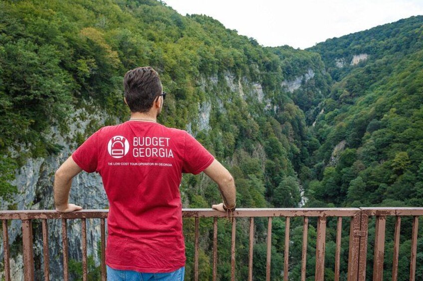 A person in a red shirt with 'BUDGET GEORGIA' on the back, standing on a wooden railing overlooking a lush green valley with steep cliffs. 2