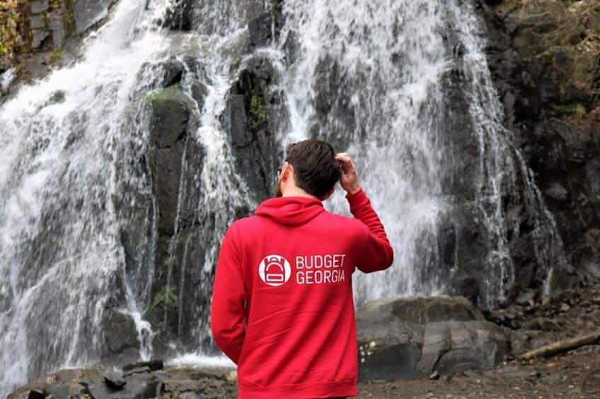 A person in a red hoodie with 'BUDGET GEORGIA' text standing in front of a waterfall. 1