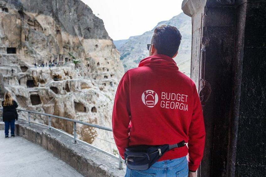 Une personne vêtue d’une veste rouge, avec l’inscription « BUDGET GEORGIA » dans le dos, se tient près d’une falaise rocheuse où se trouvent d’anciennes habitations troglodytes. 1
