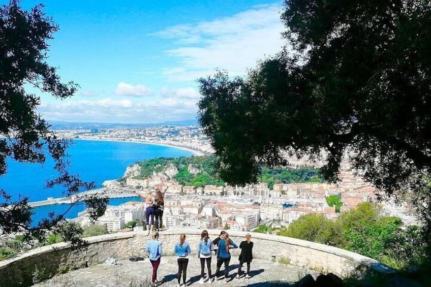 A group of people on a stone platform overlooking a coastal city with a clear blue sea and green hills in the distance. 2