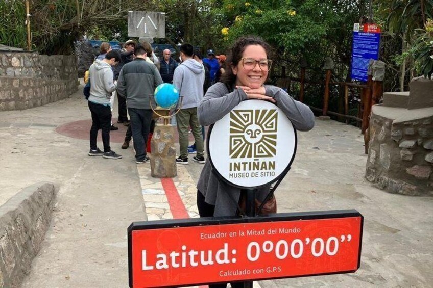 A person holding a sign with the Intitán Museum of the Site logo, a red sign with 'Latitud: 0°00'00''' and a blue informational sign in the background. 3