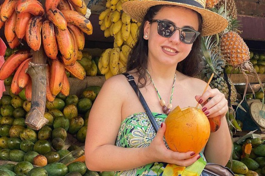 Una persona sosteniendo un coco con una pajita, rodeada de diversas frutas como plátanos y piñas. 4