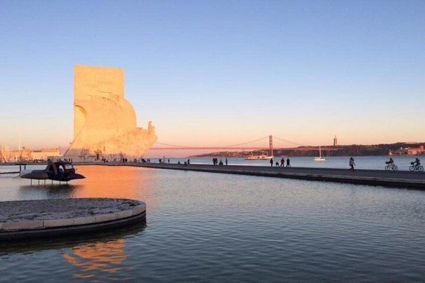 A waterfront with a large rock formation, a bridge in the background, and people walking and cycling. 1