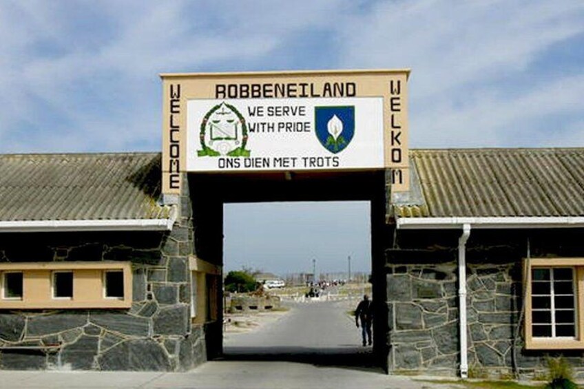 Un cartello con la scritta 'Robben Island' e 'Benvenuti', che accoglie i visitatori, con un edificio in pietra e una strada che si allontana verso l'orizzonte. 2