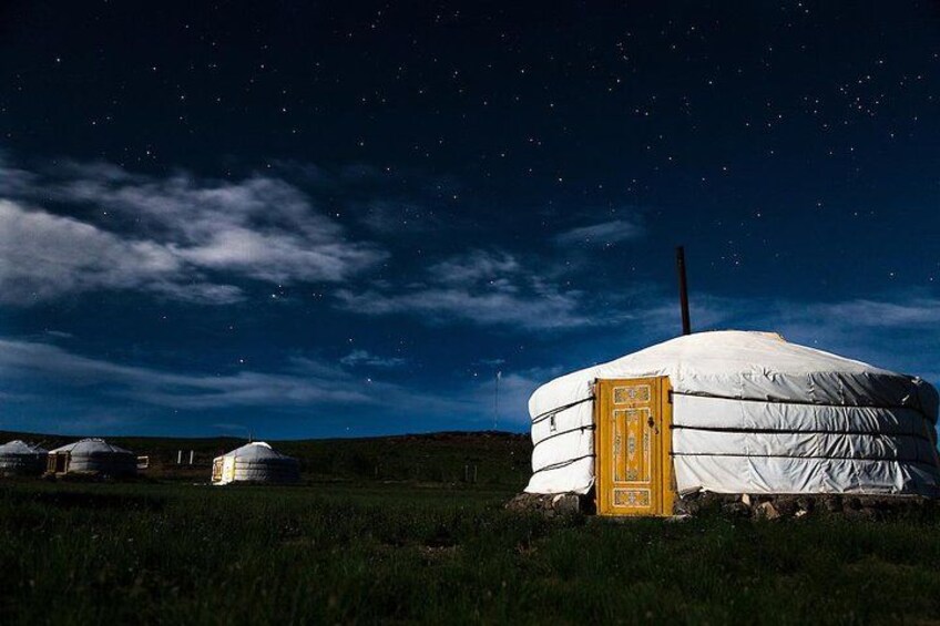 A yurt with a yellow door, surrounded by grassy terrain and other yurts under a starry night sky. 2