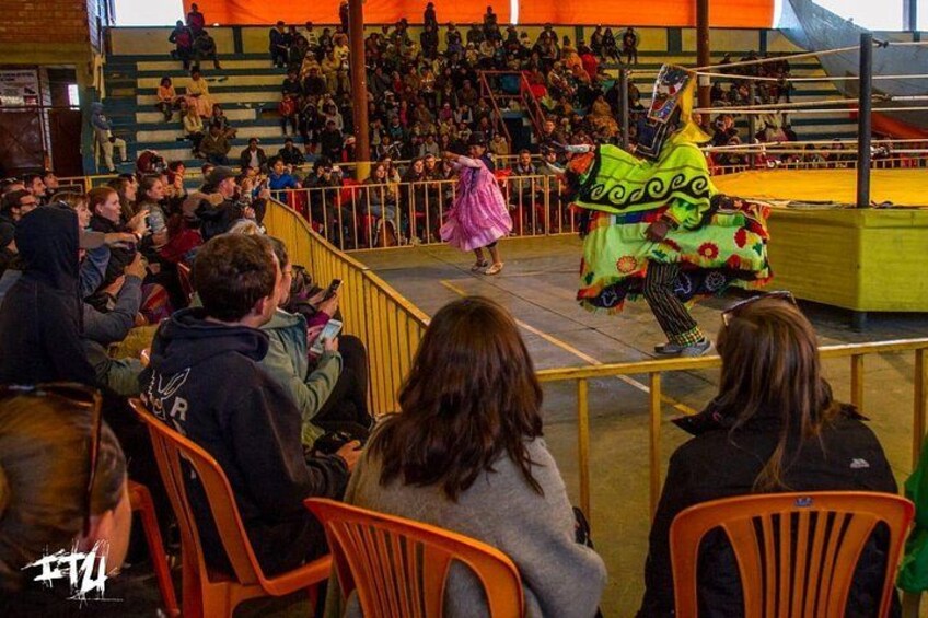 Un artista con un disfraz llamativo en el escenario, frente a un público sentado en sillas naranjas. 2