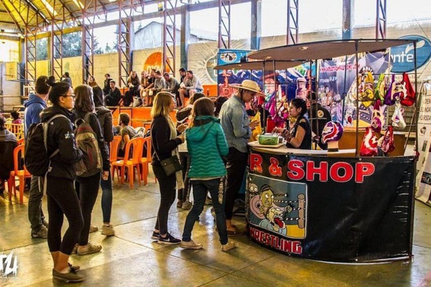 Un evento multitudinario en interior con un puesto identificado como 'R&SHOP' que vende artículos de lucha libre. 5