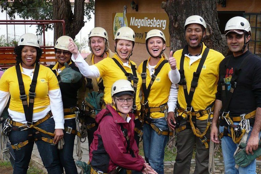 A group of people wearing helmets and harnesses, standing in front of a building with a sign that reads 'Magaliesberg.' 1