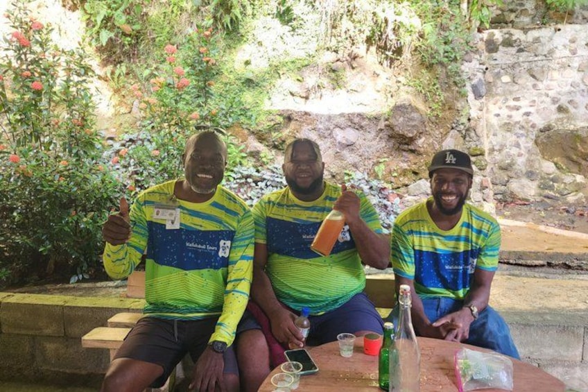 Three people sitting at a table outdoors with greenery and a stone wall in the background. 3