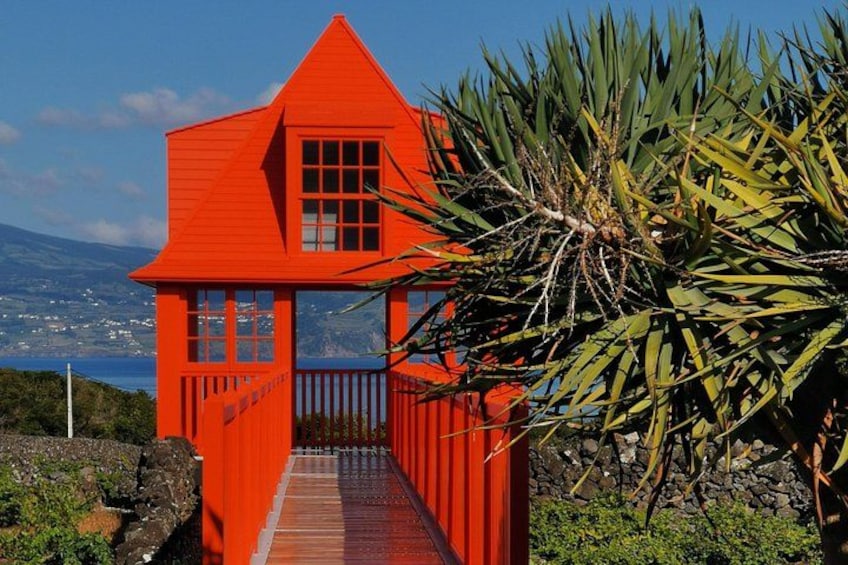 Une maison rouge avec une terrasse en bois, entourée de verdure et offrant une vue sur un paysage côtier. 3