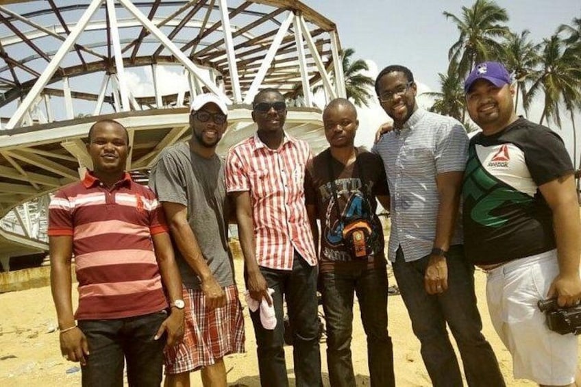 A group of people standing in front of a large, white, dome-like structure with a metal framework, palm trees in the background, and a sandy ground. 2