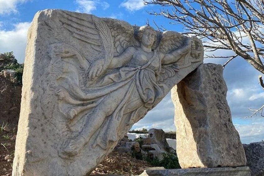 En stenskulptur af en engel med vinger, foran et klippefyldt landskab med klar himmel. 5