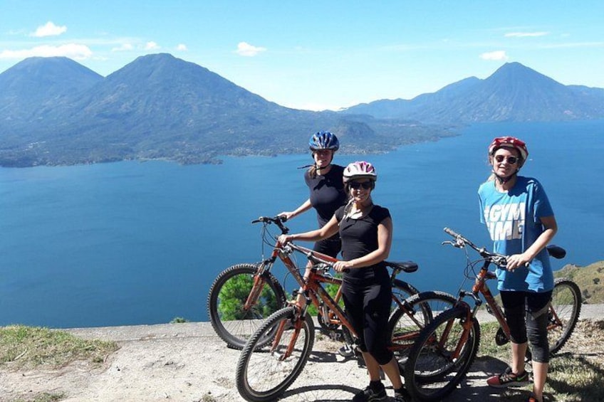 Three people with bicycles on a dirt path with a lake and mountains in the background. 1