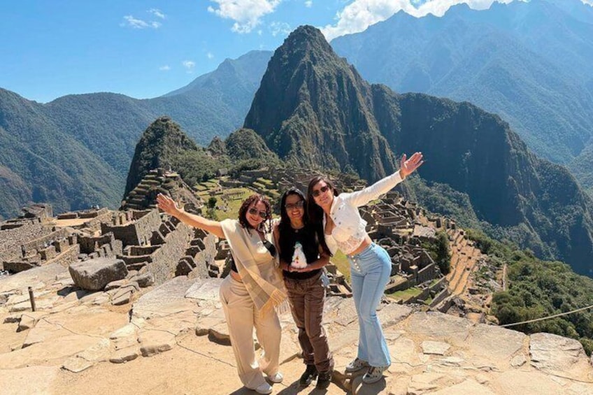 Three people standing on a stone platform with outstretched arms, overlooking ancient ruins and a mountainous landscape. 1