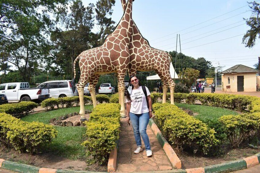A person standing in front of a giraffe statue in a landscaped area with bushes and trees. 4