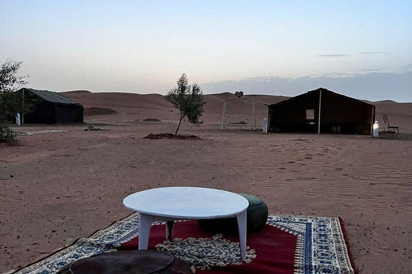 A desert landscape with a white table and red and blue patterned rug, surrounded by sand dunes and two wooden structures. 4
