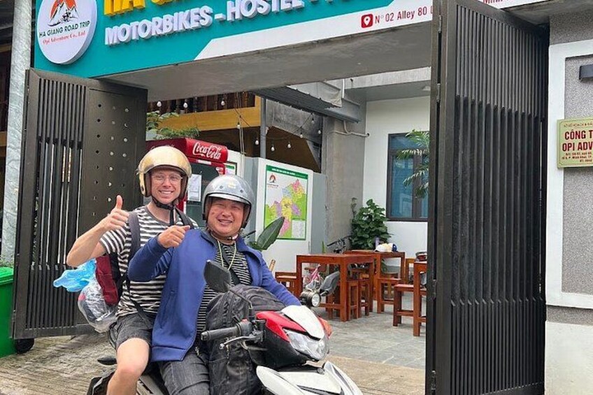 Two people on a motorcycle in front of a building with a sign that reads 'MOTORBIKES - HOSTEL'. 1
