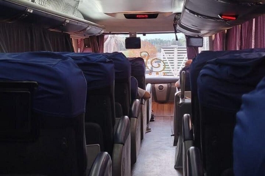 A bus interior with rows of seats, a steering wheel, and overhead luggage compartments. 3