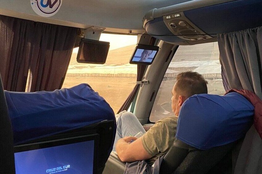 A bus interior with a man seated, a television screen displaying 'CRUZ DE SUR', and a window showing a desert landscape. 2