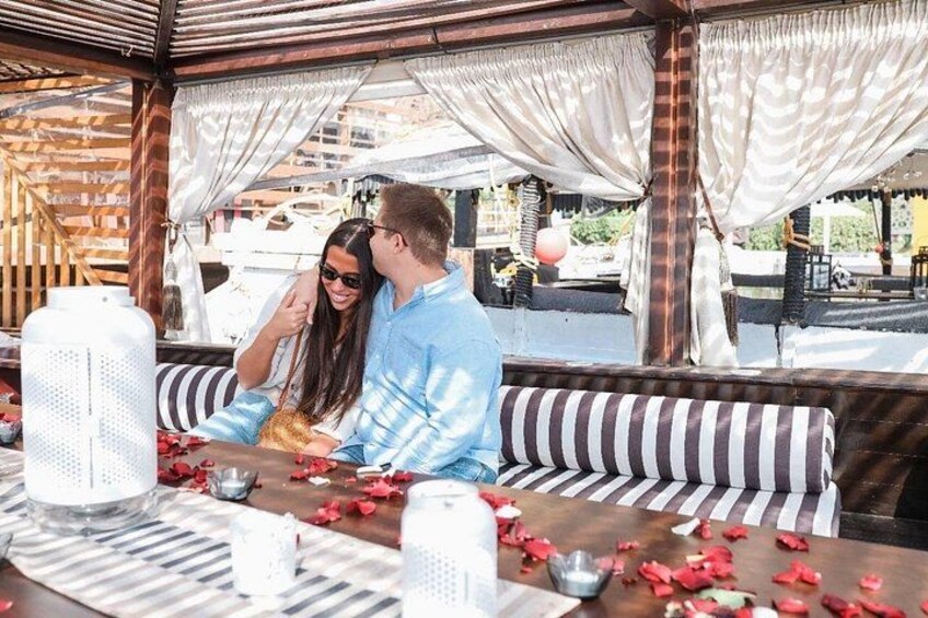 Una pareja sentada en una mesa con pétalos de rosa esparcidos en el suelo, un cojín a rayas y una jarra blanca. 5