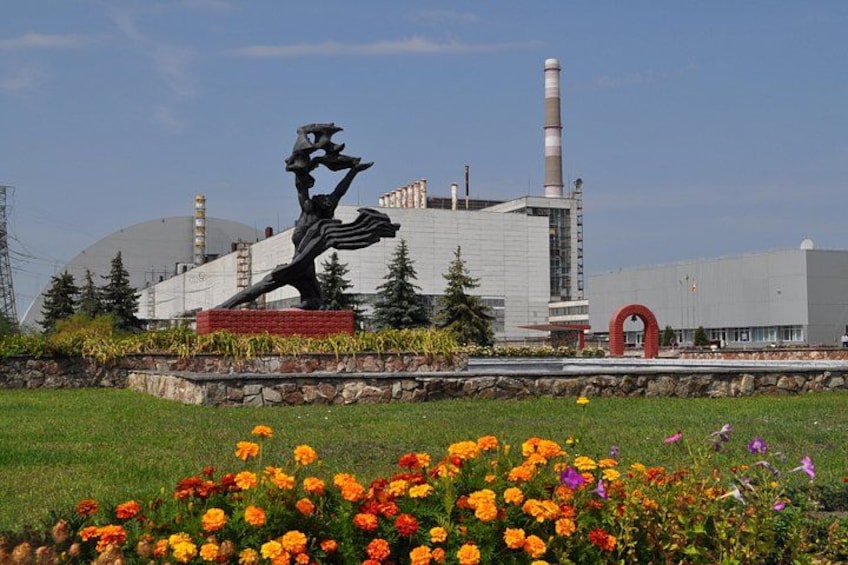 A sculpture in a park with flowers in the foreground and a large industrial building in the background. 5