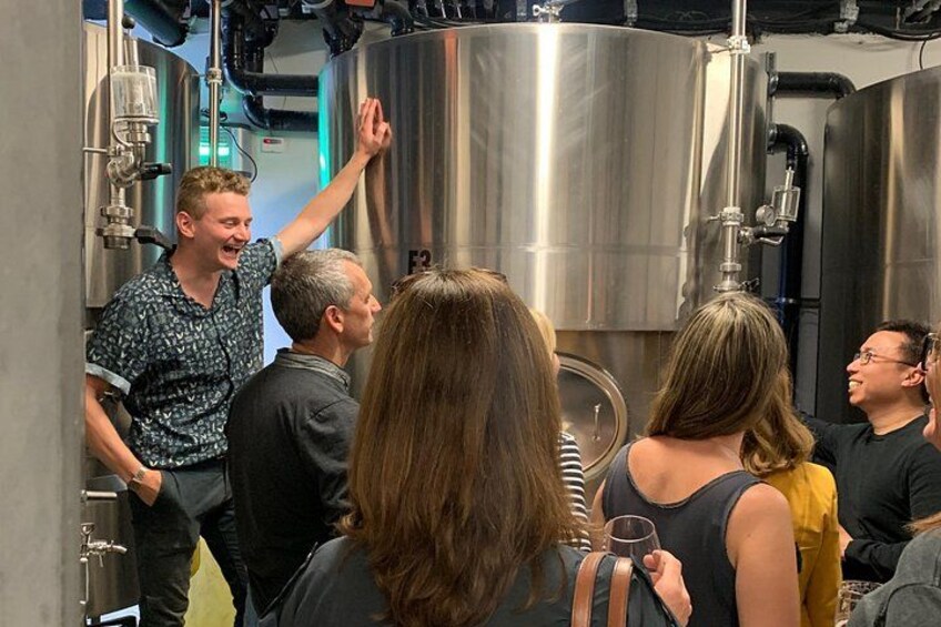 A group of people in a brewery setting with stainless steel tanks and brewing equipment. 1