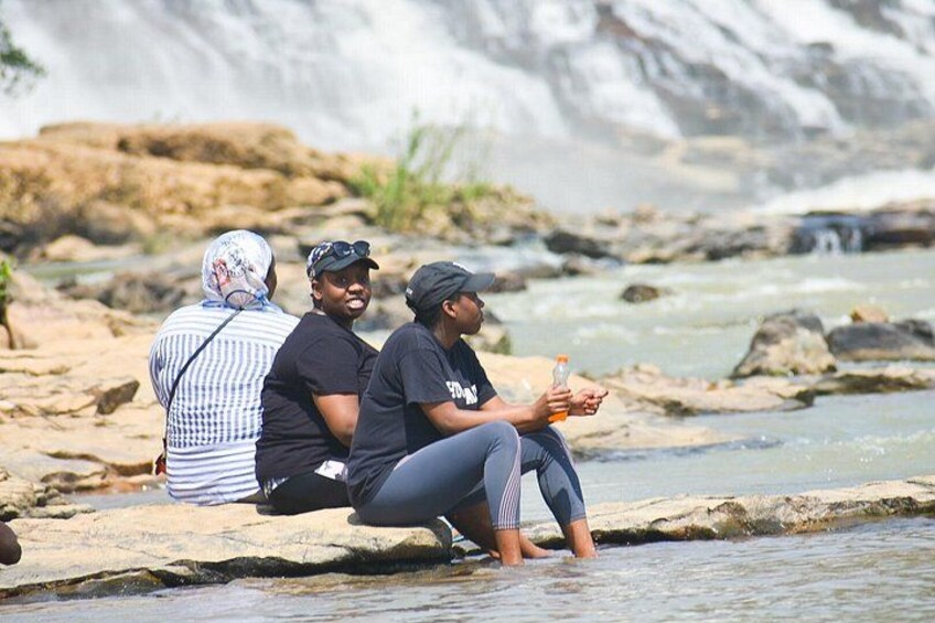 Three people sitting on rocks by a river with a waterfall in the background. 1
