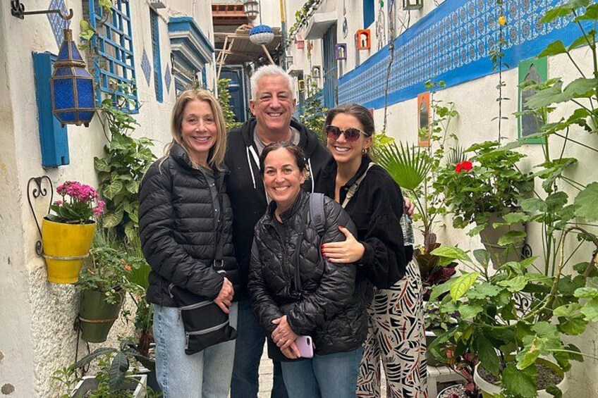 A group of people standing in a narrow street with blue and white buildings, potted plants, and a yellow flower pot. 3