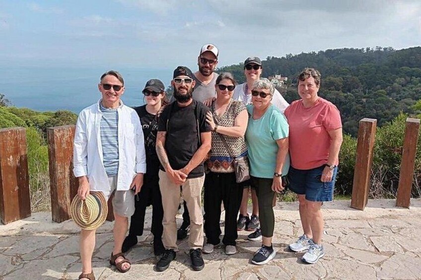 Un grupo de personas de pie sobre un sendero de piedra, con una vista panorámica de una zona boscosa y una masa de agua al fondo. 3