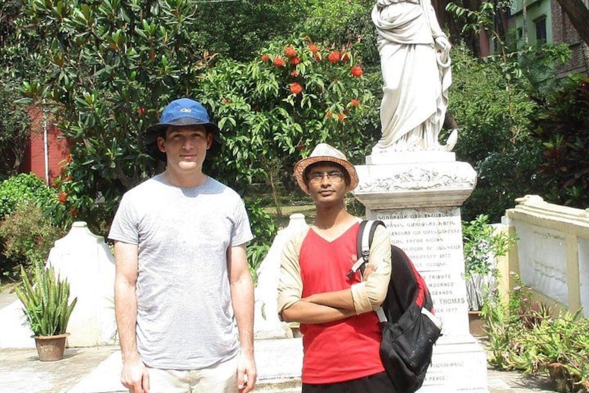 Dos personas de pie frente a una estatua con inscripciones, rodeadas de follaje y plantas en macetas. 5