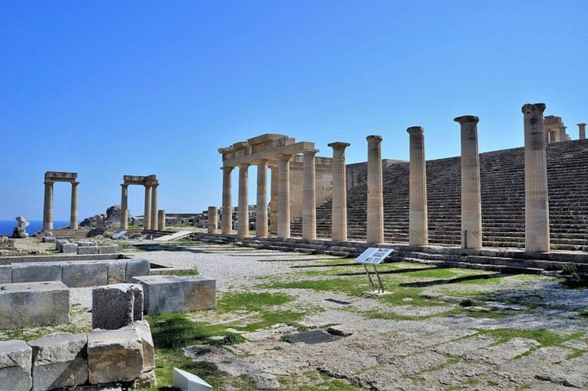 Ancient ruins with columns and stone structures, a clear sky, and a signboard. 1