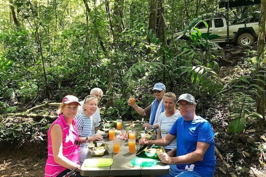 Un grupo de personas cenando al aire libre en un área boscosa, con una mesa dispuesta para una comida. 2