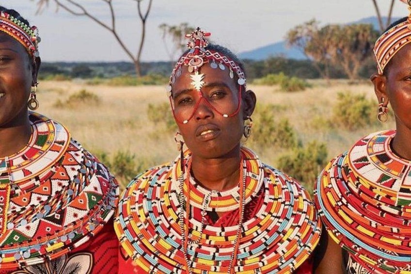 Three individuals in traditional attire with intricate beadwork and jewelry, standing in a natural landscape with sparse vegetation and distant hills. 2