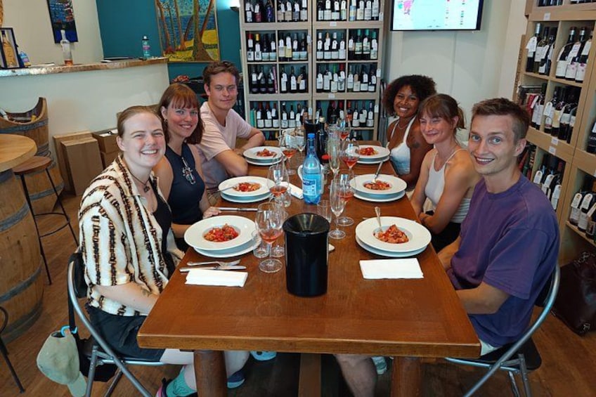 A group of people seated around a wooden table with plates of food, glasses, and bottles, in a room with shelves of bottles in the background. 5