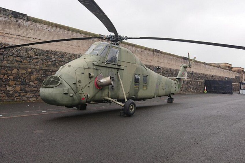 Un elicottero militare parcheggiato su una superficie asfaltata, con un muro di pietra sullo sfondo. 1