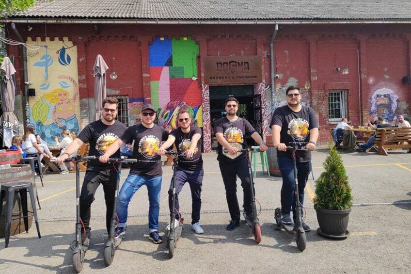Cuatro personas en bicicletas estáticas frente a un edificio con un mural y un letrero que dice 'DOGMA BREWERY & TAP ROOM'. 2