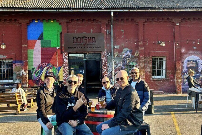 Un grupo de personas sentadas en un banco frente a un edificio con un mural colorido y un letrero que dice 'DOGMA BREWERY & TAP ROOM'. 5