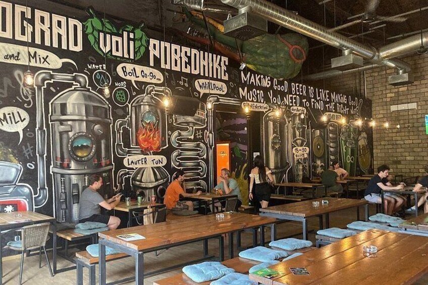 Interior de una cervecería con equipo de elaboración, mesas y clientes. 3