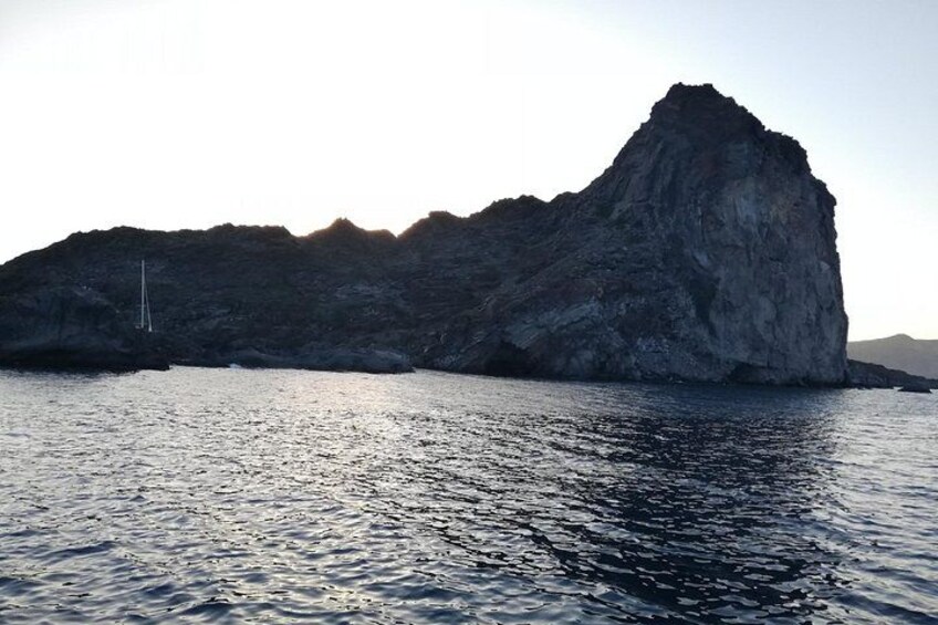 A large rock formation by the sea with calm water and a sailboat in the distance. 2