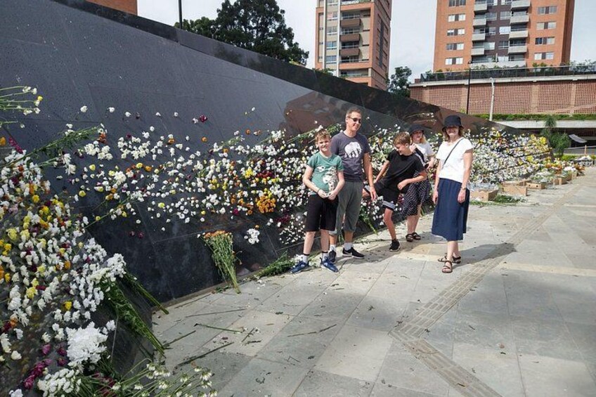 Un muro conmemorativo adornado con flores y un grupo de personas de pie frente a él. 1