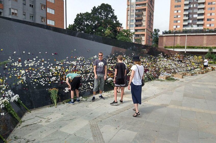 Un muro conmemorativo adornado con flores, un sendero pavimentado y edificios residenciales al fondo. 4