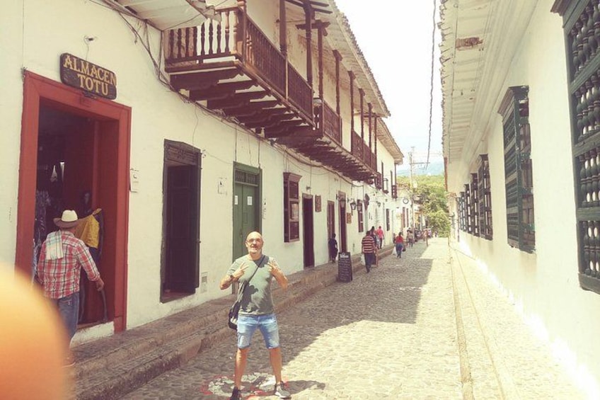 Una calle adoquinada con edificios, un hombre con camiseta y pantalones cortos, y un letrero que dice 'ALMACEN TOTO'. 2