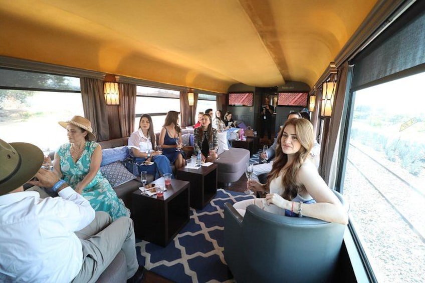A train interior with passengers seated on sofas and chairs, a table with drinks, and a window with a view of the outside. 2