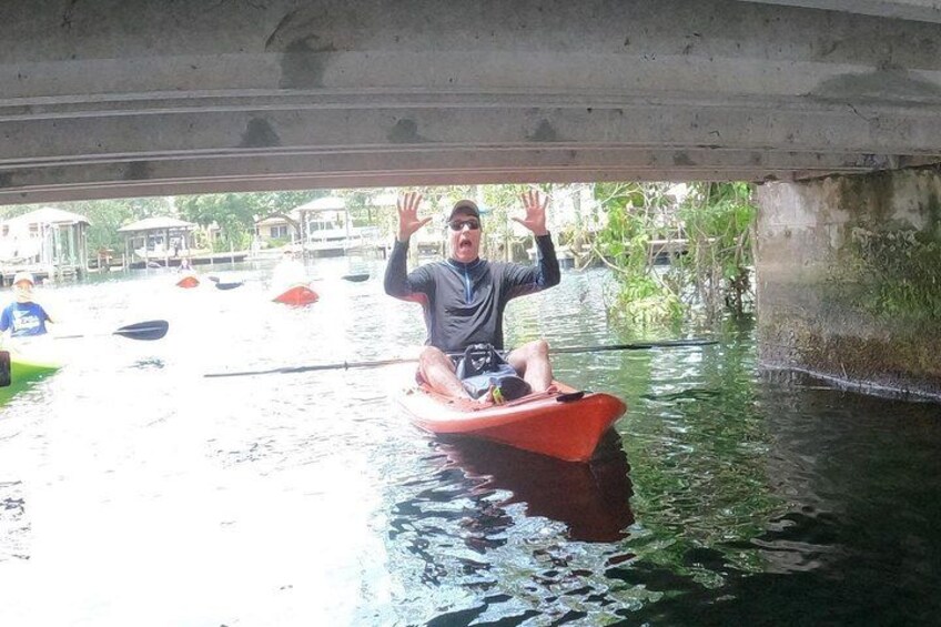 Una persona en kayak bajo un puente, rodeada de agua tranquila y vegetación. 3
