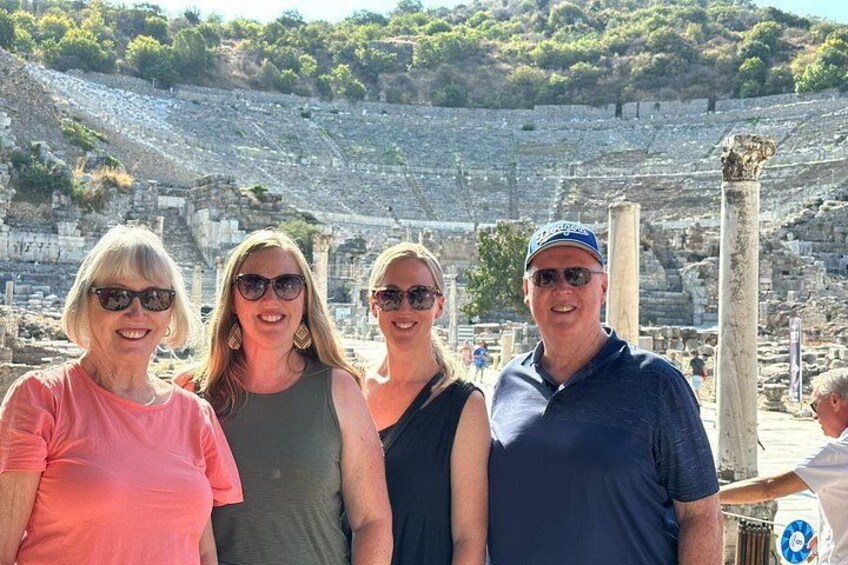 Un groupe de personnes se tient devant des ruines antiques, avec des colonnes et un grand amphithéâtre en arrière-plan. 2