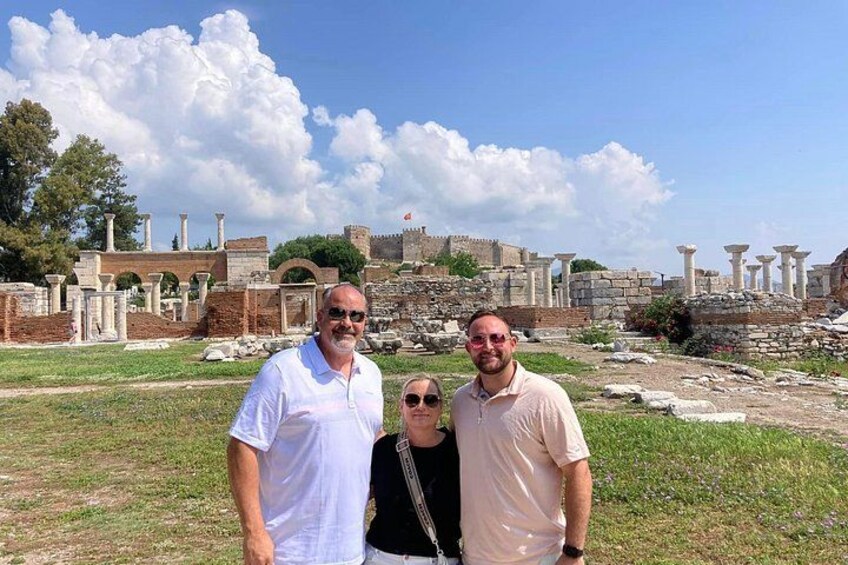 Three people standing in front of ancient ruins with columns and a fort in the background. 5