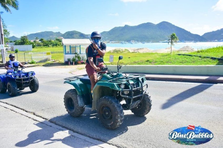 Deux personnes conduisent des quads sur une route, avec une plage et des montagnes en arrière-plan. 2