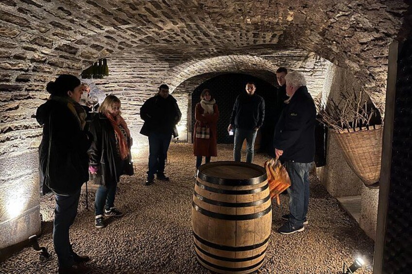 A group of people inside a stone-walled underground structure with a wooden barrel in the foreground. 2