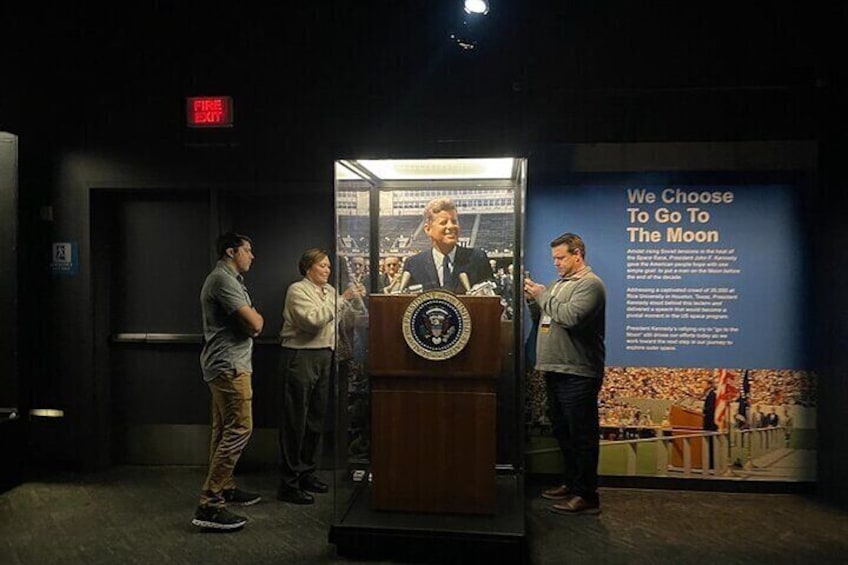 A group of people standing in front of a display with a podium and a sign that reads "We Choose To Go To The Moon". 2