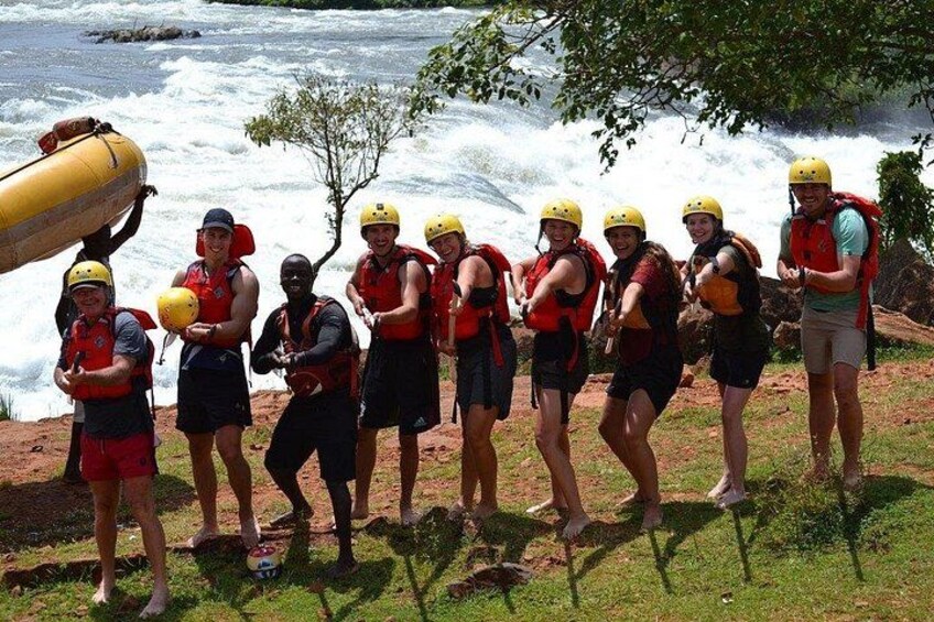 A group of people wearing helmets and life jackets, standing on a grassy area near a river with waves. 2